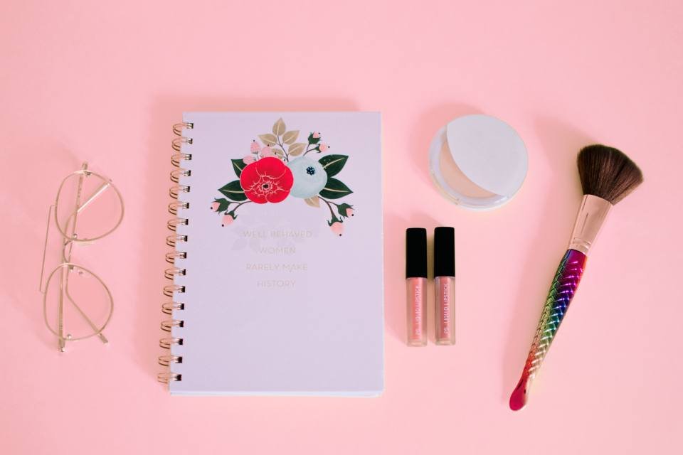 A pink flat lay with a notebook, lip gloss and brush